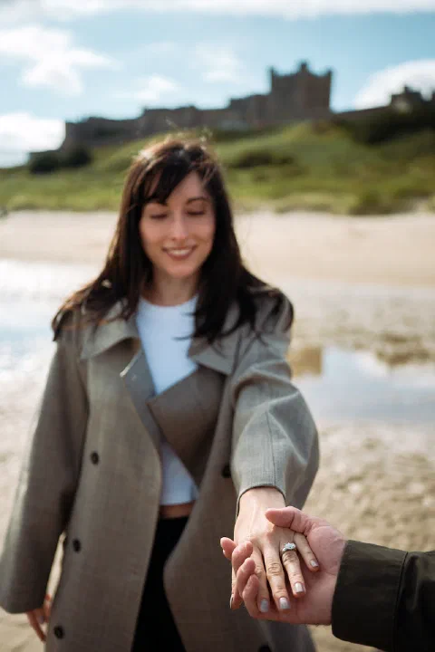 A newly engaged woman showing her ring while holding her partner's hand on Bamburgh Beach