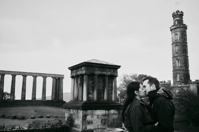 An engaged couple kissing on Calton Hill with stone monuments and a tower behind them